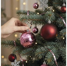 Eine Hand hängt eine glänzende rosa Weihnachtskugel an einen geschmückten Weihnachtsbaum mit dunkelroten Kugeln und Lichtern.