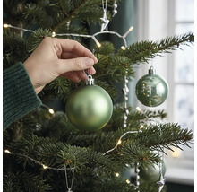 Eine Hand hängt eine matte grüne Weihnachtskugel an einen geschmückten Weihnachtsbaum mit LED Lichterkette.