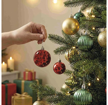 Eine Hand hängt eine rote Weihnachtskugel an einen geschmückten Weihnachtsbaum mit Lichtern und Geschenken im Hintergrund.