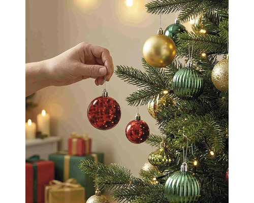 Eine Hand hängt eine rote Weihnachtskugel an einen geschmückten Weihnachtsbaum mit Lichtern und Geschenken im Hintergrund.