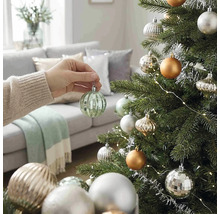 Eine Hand hängt eine gerippte grüne Weihnachtskugel aus Glas an einen geschmückten Weihnachtsbaum in einem hellen Wohnzimmer.