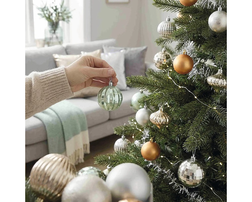 Eine Hand hängt eine gerippte grüne Weihnachtskugel aus Glas an einen geschmückten Weihnachtsbaum in einem hellen Wohnzimmer.