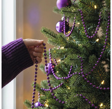 Eine Hand dekoriert einen Weihnachtsbaum mit einer lila Perlengirlande, Christbaumkugeln und warmweißer Beleuchtung.