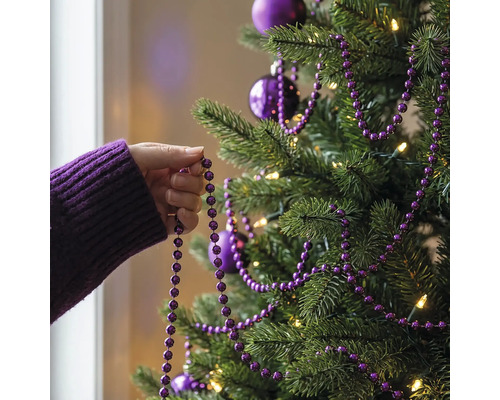 Eine Hand dekoriert einen Weihnachtsbaum mit einer lila Perlengirlande, Christbaumkugeln und warmweißer Beleuchtung.