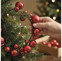 Hand beim Dekorieren eines Weihnachtsbaums mit einer Girlande aus roten Weihnachtskugeln verschiedener Grössen und warmweissen Lichtern.