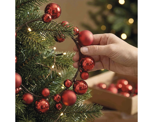 Hand beim Dekorieren eines Weihnachtsbaums mit einer Girlande aus roten Weihnachtskugeln verschiedener Grössen und warmweissen Lichtern.