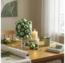 Festliche Tischdekoration mit einer Glasvase voller grüner Weihnachtskugeln, einer brennenden Kerze und einer Tasse auf einem Holztisch.