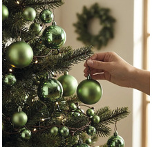 Hand hängt eine grüne matte Kugel an einen geschmückten Weihnachtsbaum mit glänzenden und matten Kugeln sowie Lichtern.