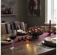 Festlich gedeckter Holztisch mit Tischläufer, Schale voll mit Weihnachtskugeln und Lichterkette, Weinglas und Tellern in elegantem Ambiente.