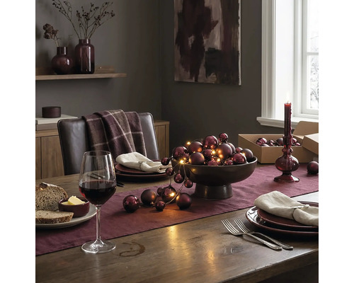 Festlich gedeckter Holztisch mit Tischläufer, Schale voll mit Weihnachtskugeln und Lichterkette, Weinglas und Tellern in elegantem Ambiente.