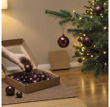 Hand entnimmt eine Weihnachtskugel aus einem Karton neben einem geschmückten Weihnachtsbaum mit Beleuchtung.