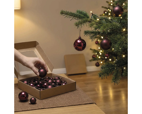 Hand entnimmt eine Weihnachtskugel aus einem Karton neben einem geschmückten Weihnachtsbaum mit Beleuchtung.