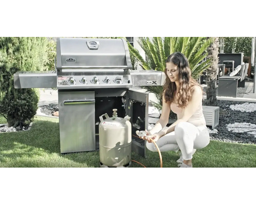 Une femme branche une bouteille de gaz sur un barbecue à gaz Napoleon en acier inoxydable dans un jardin lumineux et organisé.