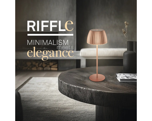 Moderne Tischleuchte Riffle mit plissiertem Schirm auf einem runden Standfuß in einem eleganten Wohnraum mit Holztisch und Kamin.