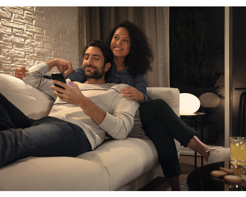 Paar entspannt auf einem Sofa in einem hellen Wohnzimmer, Mann schaut auf sein Smartphone