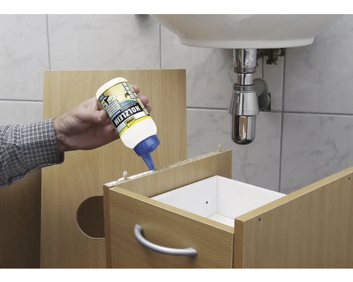 Ein Handwerker trägt Holzleim auf ein Holzmöbelstück im Badezimmer auf. Im Hintergrund befindet sich ein Waschbecken.