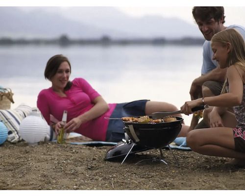 Une famille fait des grillades au bord d''un lac avec un barbecue au charbon de bois, tandis que la mère se détend avec une boisson.