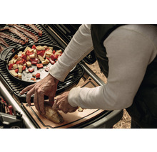 Eine Person bereitet auf einem Grill Speisen zu, darunter Speck, Gemüse in einer Pfanne und geschnittene Kartoffeln auf einem Schneidebrett.