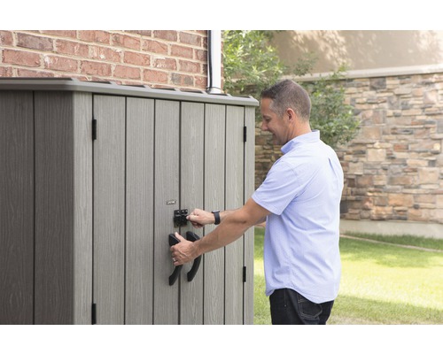 Mann öffnet eine graue Gartengerätebox mit Griffen und Schloss vor einer Ziegelwand.