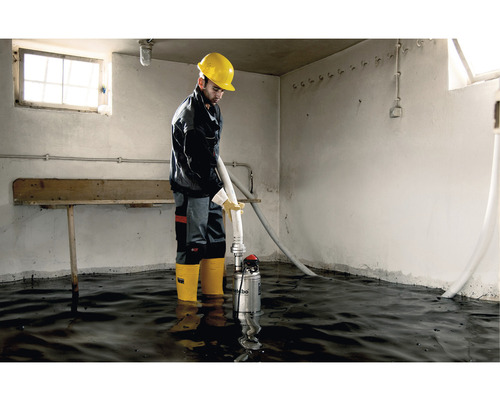 Un ouvrier avec un casque de protection jaune utilise une pompe à eau sale dans une pièce inondée.