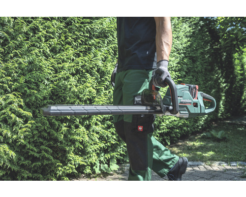 Une personne porte un taille-haie à batterie Metabo devant une haie verte