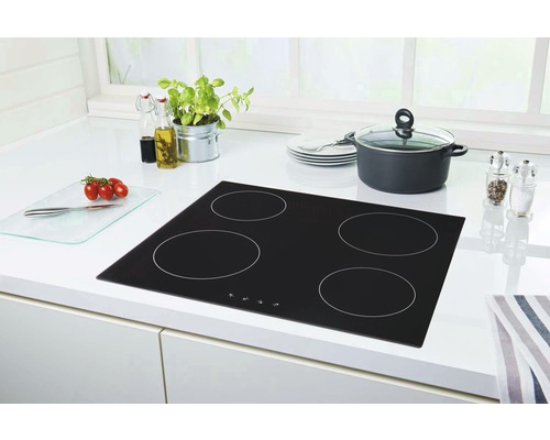 Plaque de cuisson vitrocéramique noire avec quatre zones de cuisson sur un plan de travail de cuisine blanc avec une marmite, une pile d''assiettes, un moulin à épices, une planche à découper, des tomates et des herbes.