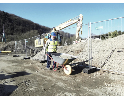 Ouvrier pousse une brouette avec des déchets de construction devant une clôture de chantier et une excavatrice.