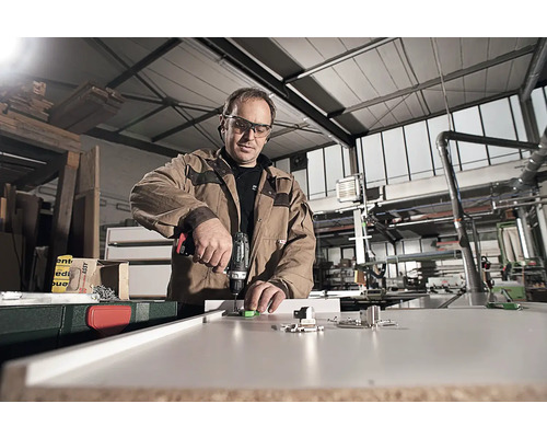 Un homme monte des ferrures sur un meuble avec une perceuse dans un atelier.