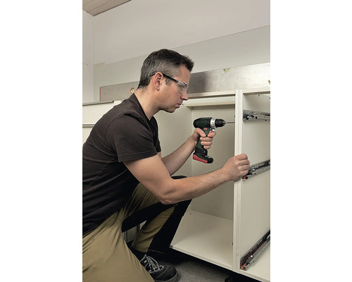 Un homme installe des coulisses de tiroir avec une visseuse sans fil sur un meuble de cuisine.