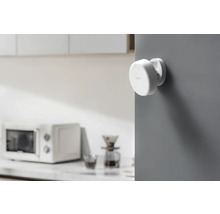 Un capteur de porte et de fenêtre Aqara blanc est monté sur un mur gris. Un four à micro-ondes se trouve à l''arrière-plan.