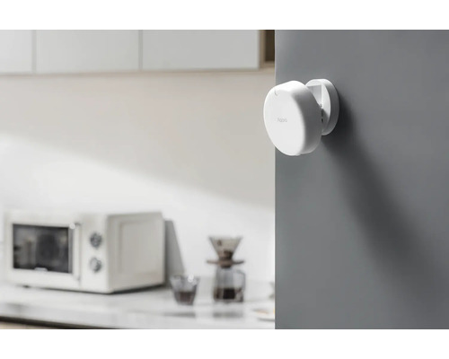 Un capteur de porte et de fenêtre Aqara blanc est monté sur un mur gris. Un four à micro-ondes se trouve à l''arrière-plan.