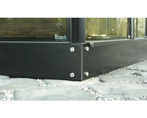 Vue détaillée d''une fondation de serre en métal avec fixation par vis sur un sol en dalles de pierre. Logo Vitavia.