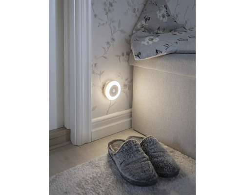 Scène avec une veilleuse ronde à LED avec détecteur de mouvement fixée au mur, des chaussons et un tapis