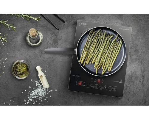 Plaque à induction avec asperges dans une poêle, ainsi que des ingrédients tels que de l''huile et des épices.