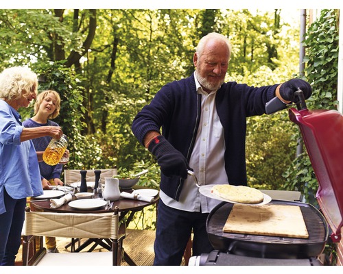 Im Freien bereitet eine Familie Pizza auf einem Grill zu. Ein gedeckter Tisch steht bereit.