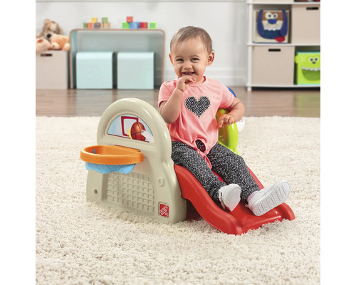 Un enfant est assis sur un toboggan Step2 avec un panier de basket dans une salle de jeux.