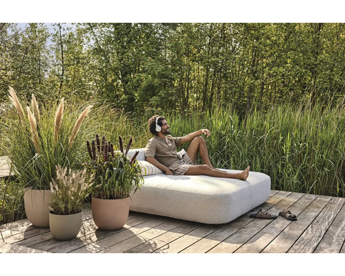 Scène avec un homme portant un casque audio se détendant sur un lit de jour dans le jardin, entouré de pots de fleurs plantés.