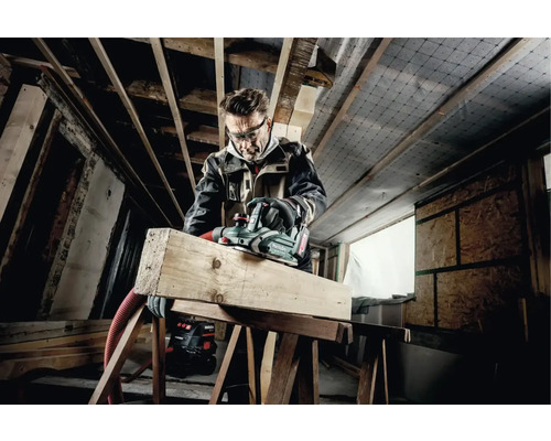 Un artisan portant des lunettes de protection rabote une planche de bois avec un rabot électrique Metabo dans une construction brute.