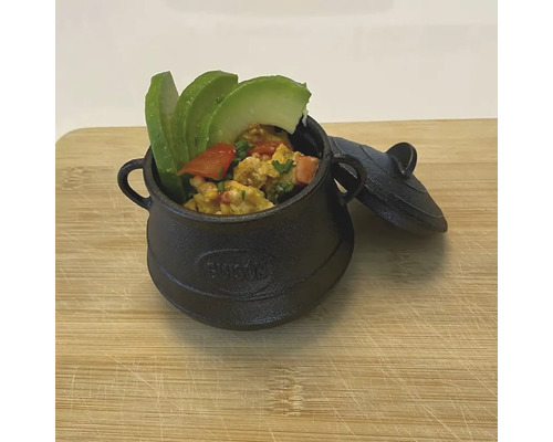Schwarzer Mini-Gusseisentopf mit Essen und Deckel auf einem Schneidebrett