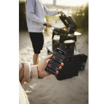 Eine Person grilliert mit einem Grill und kontrolliert die Temperatur mit einer App auf einem Smartphone.