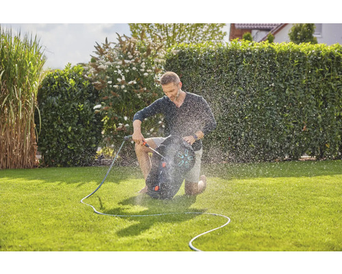 Ein Mann auf einem Rasen kniet und eine Gartenschlauchbox bedient, während Wasser versprüht wird.