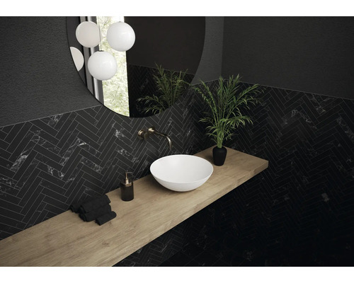 Badezimmer mit Waschbecken, Spiegel, Armatur, Pflanze und Handtüchern auf einer Holzablage vor einer Wand mit Fliesen im Fischgrätmuster.