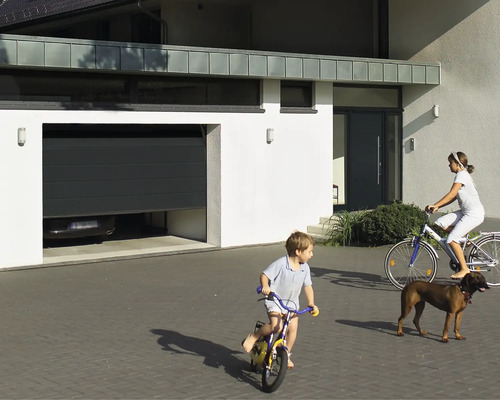 Porte sectionnelle Hörmann EcoStarMatic anthracite RAL 7016 avec rainures M 2375x2125 mm avec motorisation de porte de garage Liftronic 700 2e génération Scène avec maison, porte de garage, porte d''entrée, enfants qui jouent et un chien.