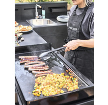 Une femme prépare des saucisses et des légumes sur un barbecue dans une cuisine extérieure moderne.