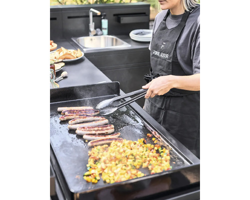 Une femme prépare des saucisses et des légumes sur un barbecue dans une cuisine extérieure moderne.