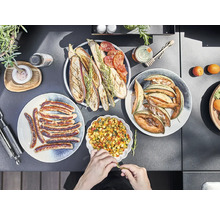 Divers plats dans des assiettes, notamment des saucisses, des légumes grillés, du melon et de la salade, sur un plateau de table sombre