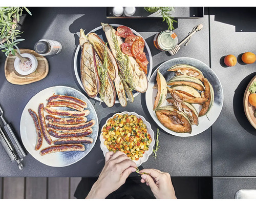 Divers plats dans des assiettes, notamment des saucisses, des légumes grillés, du melon et de la salade, sur un plateau de table sombre