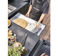 Szene einer Person mit Grillschürze, die eine Schublade mit Grillutensilien wie Topflappen, Wender und Serviergabel öffnet, auf einem Tresen stehen außerdem eine Platte mit gegrilltem Gemüse, eine Flasche Olivenöl, Gewürze und eine Zange