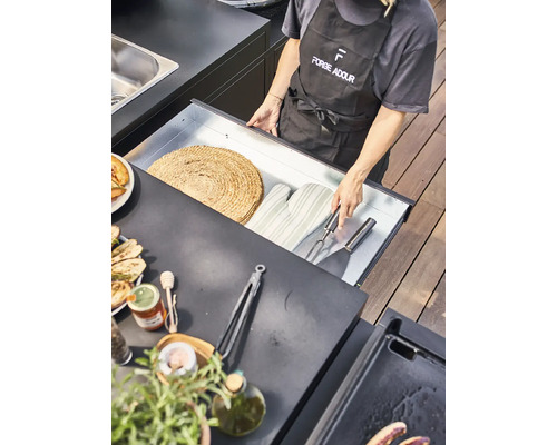 Szene einer Person mit Grillschürze, die eine Schublade mit Grillutensilien wie Topflappen, Wender und Serviergabel öffnet, auf einem Tresen stehen außerdem eine Platte mit gegrilltem Gemüse, eine Flasche Olivenöl, Gewürze und eine Zange