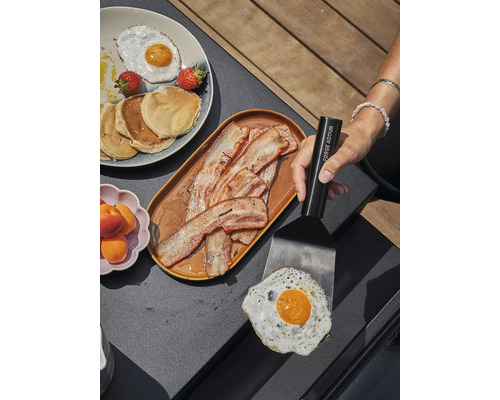 Scène de petit-déjeuner avec œuf au plat, bacon, crêpes et fruits sur une table, une spatule tenant un œuf au plat.